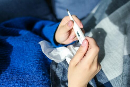 A woman checks a digital thermometer while lying indoors, showing signs of illness.